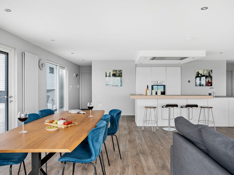 A dining area with a wooden table set with food and drinks and blue chairs next to a kitchen with white cabinets and bar stools at Huxham View and Annex in Stoke Canon