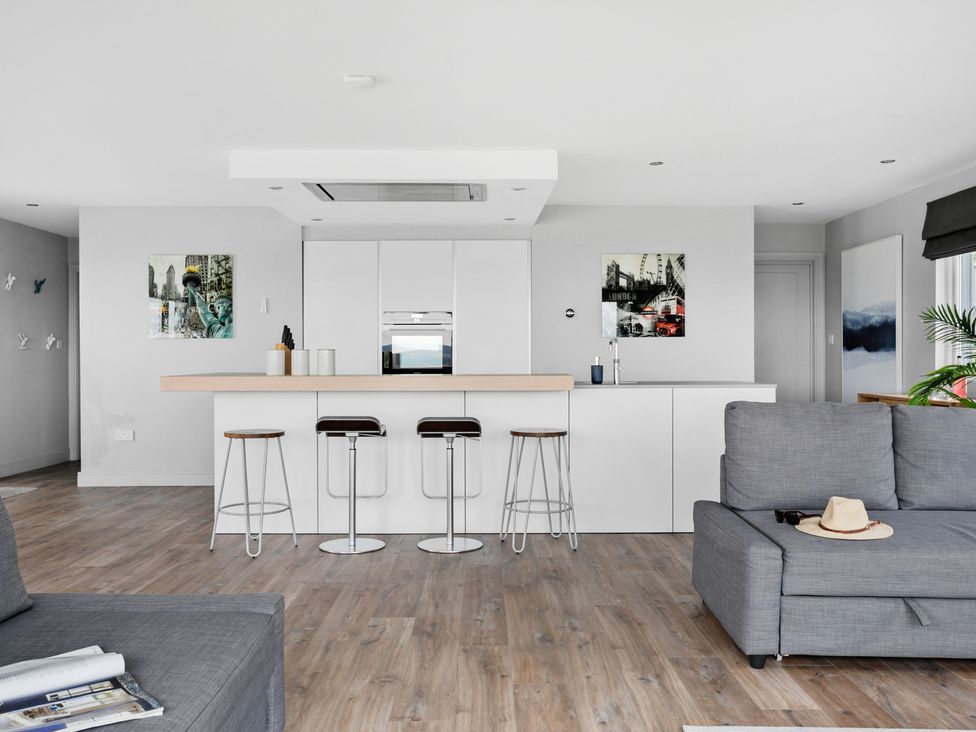 A modern kitchen with white cabinets, a wooden countertop, four bar stools, a gray sofa with a hat and sunglasses, and wall art at Huxham View and Annex in Stoke Canon