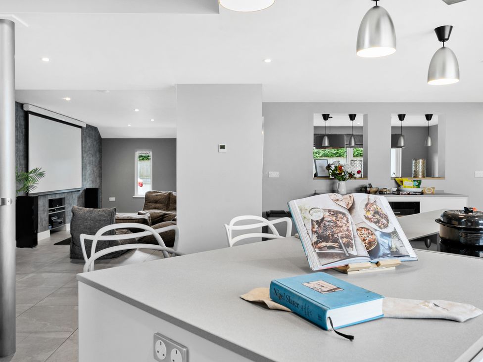 A kitchen island with cookbooks and modern pendant lights overlooking a living area with sofas and a projector screen at Huxham View and Annex in Stoke Canon