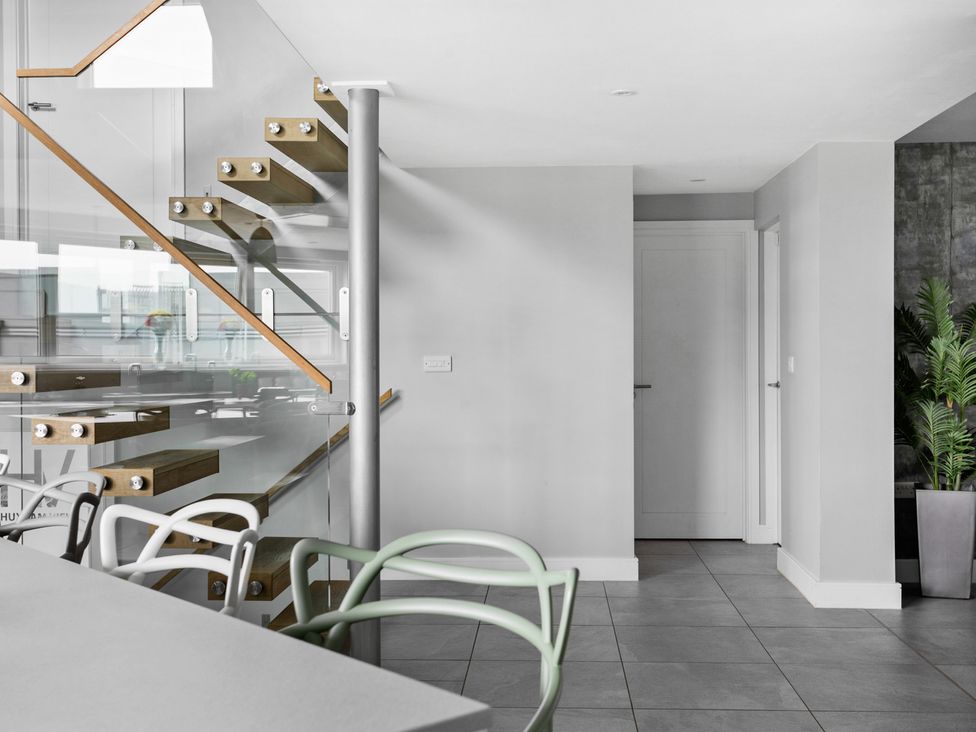 A modern interior with floating wooden stairs and glass railing a dining area with contemporary chairs and a white door at Huxham View and Annex in Stoke Canon