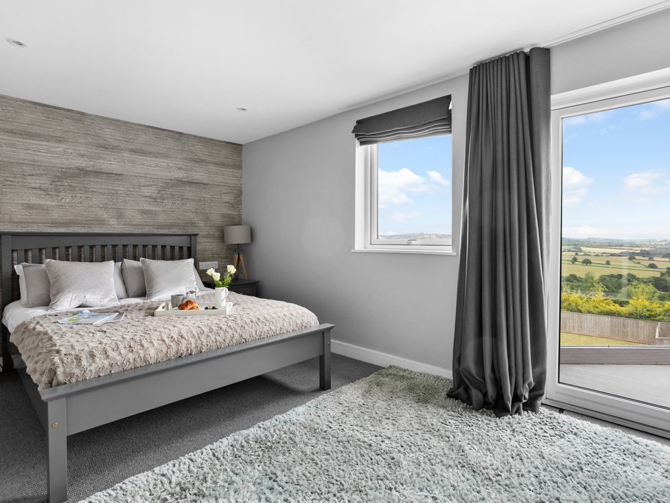A bedroom with a bed a bedside table a lamp window with curtains and a glass door showing a landscape at Huxham View and Annex in Stoke Canon